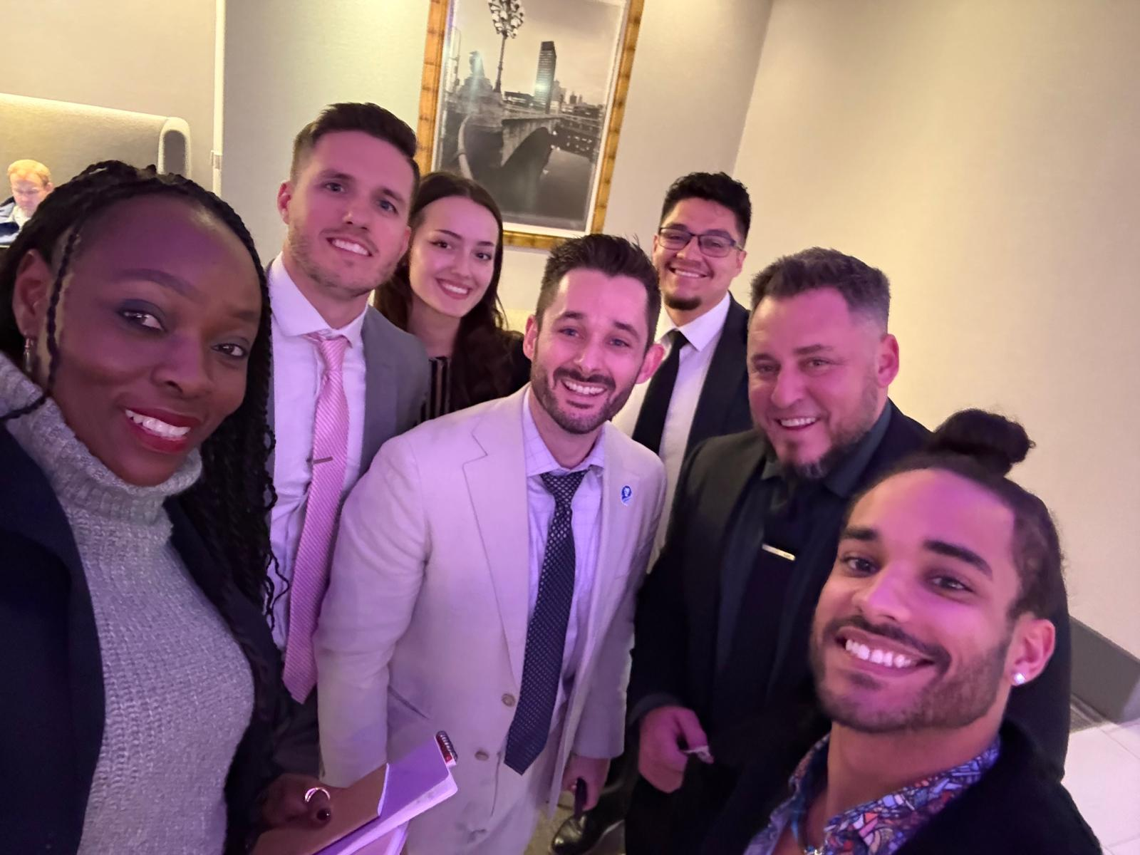 Seven professionally dressed colleagues smiling together for a group selfie at an indoor event.